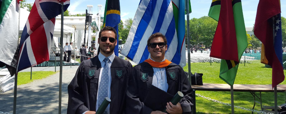 Santiago Haretche y Federico Mazzoli vestidos con toga. Detrás, la bandera uruguaya junto a otras banderas.